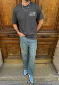 Γκρι βαμβακερό t-shirt με λογότυπο "Ford Mustang", σε συνδυασμό με ανοιχτό μπλε τζιν παντελόνι και μπλε αθλητικά παπούτσια. Ξύλινο φόντο με λαβές πορτών ορατές.