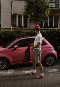 Fiatal férfi piros sapkában és rózsaszín cipőkben áll egy parkoló rózsaszín kisméretű autónál, kezében egy sötét dzsekivel egy városi utcán.