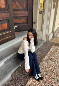 Une mannequin assise sur des marches en pierre porte un pull côtelé crème décolleté, un jean évasé bleu et des chaussures noires avec des rayures blanches.