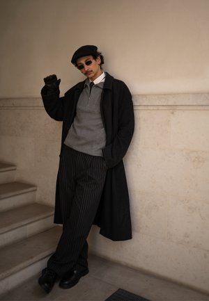 Hombre con boina negra, gafas de sol, abrigo largo negro, suéter gris y pantalones de rayas, apoyado contra una pared beige junto a unas escaleras.