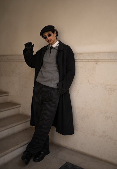 Hombre con boina negra, gafas de sol, abrigo largo negro, suéter gris y pantalones de rayas, apoyado contra una pared beige junto a unas escaleras.