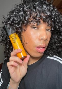 KMS Curl Up Perfecting Lotion w przezroczystej pomarańczowej butelce, z czarną pompką. Etykieta zawiera czarny tekst, określający jej zastosowanie dla kręconych włosów.
