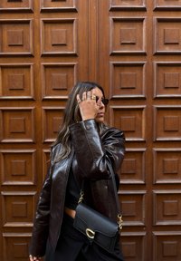 Chaqueta de cuero marrón con diseño corto, bolso negro con herrajes dorados y gafas de sol rectangulares sobre un fondo de madera texturizada.