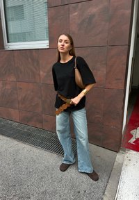 Črno oversize majico, svetlo modre široke kavbojke, rjav krožni pas, rjav ženski torbica, rjave gležnjarje, ob tem teksturiranem temnem zidu.
