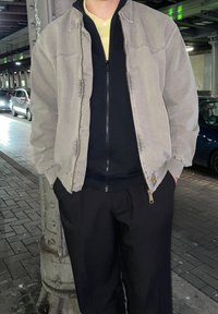 Šviesiai pilkas zip-up bomber tipo švarkas ant juodos zip-up džemperio, derinamas su laisvais juodais kelnais. Gatvės aplinka su automobiliais fone.