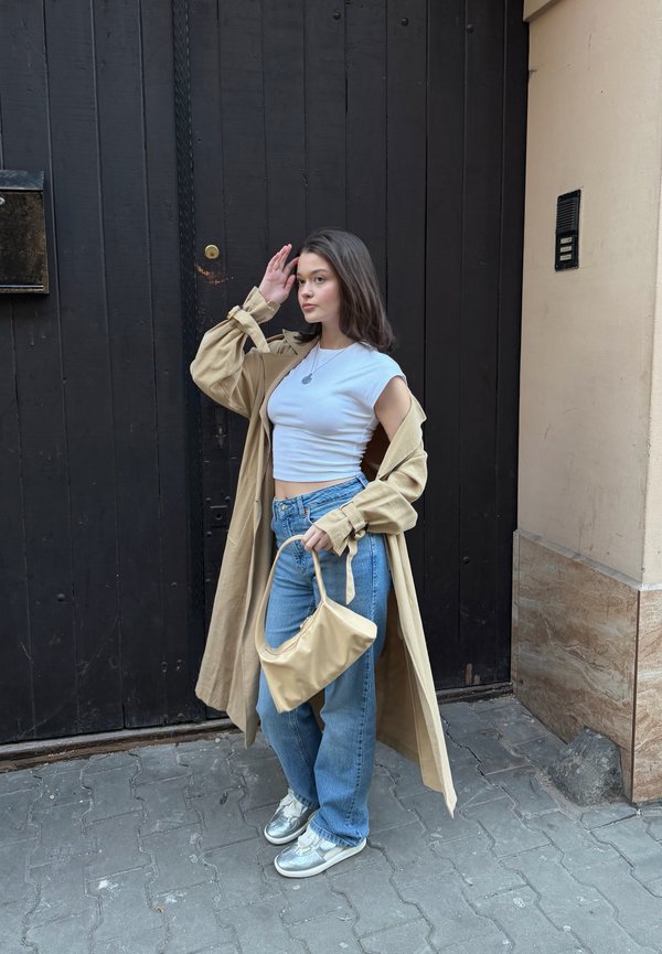 Ung kvinde iført beige trenchcoat, hvid cropped top, blå jeans, sølv sneakers, holder beige håndtaske, står på fortov ved en sort dør.