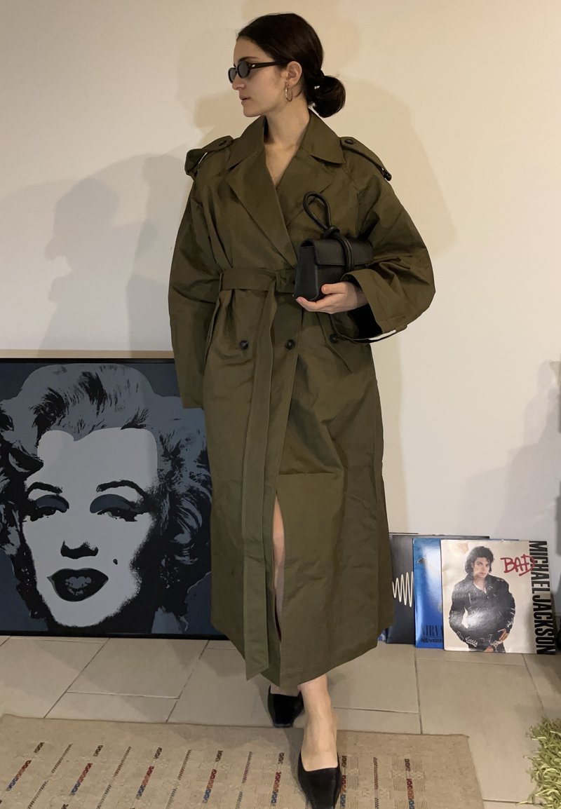 Femme en trench-coat vert olive, talons noirs, lunettes de soleil et petit sac à main noir, debout à l'intérieur près d'une œuvre emblématique de Marilyn Monroe et de disques vinyles.