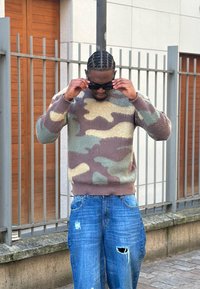 Pull en tricot avec motif camouflage aux couleurs beige, vert et brun ; associé à un jean bleu déchiré. Le mannequin ajuste ses lunettes de soleil foncées.