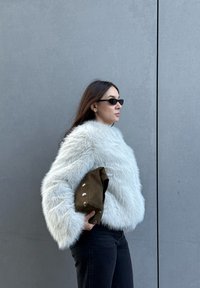 Femme portant un pull blanc duveteux et des lunettes de soleil noires, tenant une pochette marron contre un mur gris.