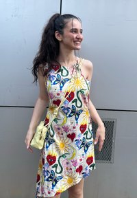 Farbiger ärmelloser Kleid mit verspielten Blumen- und Gesichtsdrucken, cremefarbenem Hintergrund und asymmetrischem Saum. Ergänzt durch eine kleine gelbe Tasche.