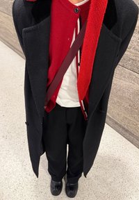 Manteau en laine noire porté sur un cardigan rouge en maille, chemise blanche en dessous, pantalon noir et chaussures noires. Comprend un détail de sangle bordeaux.