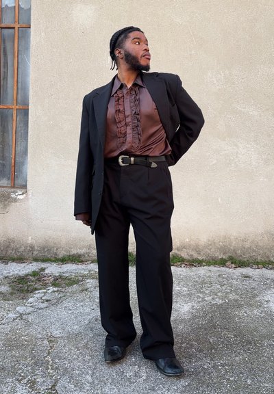 Traje negro con cierre de dos botones, combinado con una camisa marrón chocolate con volantes. Cinturón negro con acentuación en plata y zapatos negros.