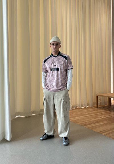 Jersey rosa claro con patrón geométrico, logo negro y acentos verdes, combinado con pantalones cargo beige y zapatos negros. Camiseta blanca visible.