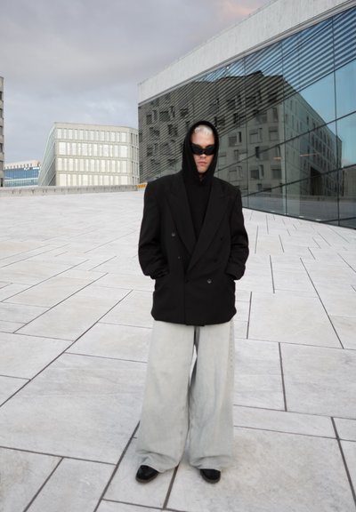 Blazer negro oversized de doble botonadura sobre una sudadera negra, combinado con jeans de pierna ancha gris claro y zapatos negros. Las gafas de sol completan el look.