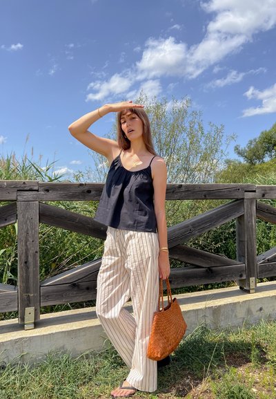 Top negro sin mangas, pantalones de rayas de pierna ancha y un bolso marrón de cuerda. Entorno al aire libre con una cerca de madera y follaje verde. Cielo brillante.
