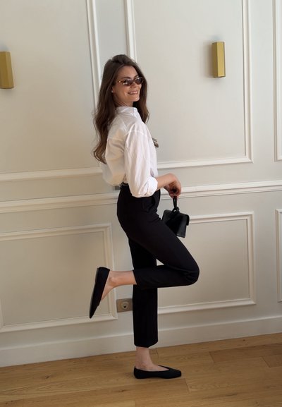 Mujer con gafas de sol, camisa blanca, pantalones negros y zapatos planos, sosteniendo un pequeño bolso negro, de pie sobre una pierna en el interior contra una pared blanca.