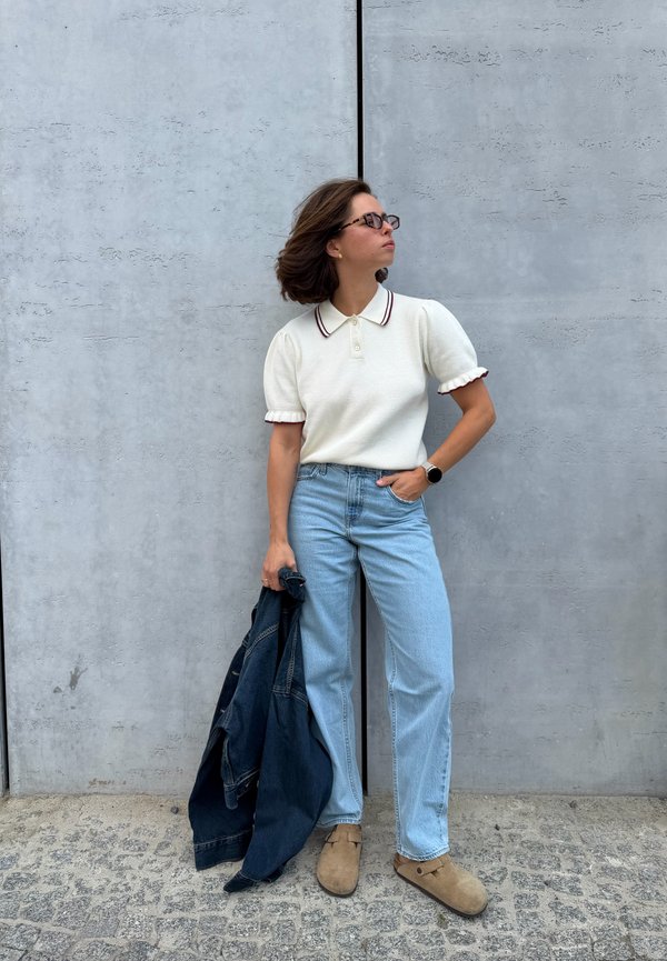 Femme portant une chemise blanche à manches courtes avec col, jean bleu clair, chaussures marron clair et lunettes, se tenant contre un mur gris en tenant une veste sombre d'une main.