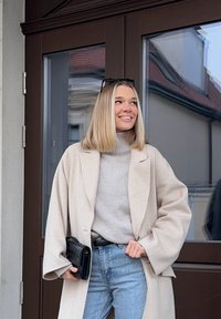 Femme aux cheveux blonds portant un manteau beige, un col roulé gris clair, un jean bleu, tenant une pochette noire, debout près d'une porte brune, souriante et regardant vers la gauche.