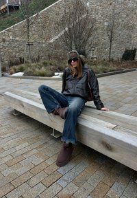 Personne portant un chapeau en fourrure, des lunettes de soleil, une veste en cuir noire, un jean bleu et des bottes marron, assise sur un banc en bois à l'extérieur près de murs en pierre.