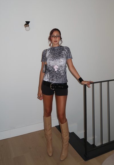 Mujer con top de estampado de leopardo, pantalones cortos negros, cinturón ancho, botas beige hasta la rodilla, gafas y joyería, de pie en el interior cerca de la barandilla de una escalera.