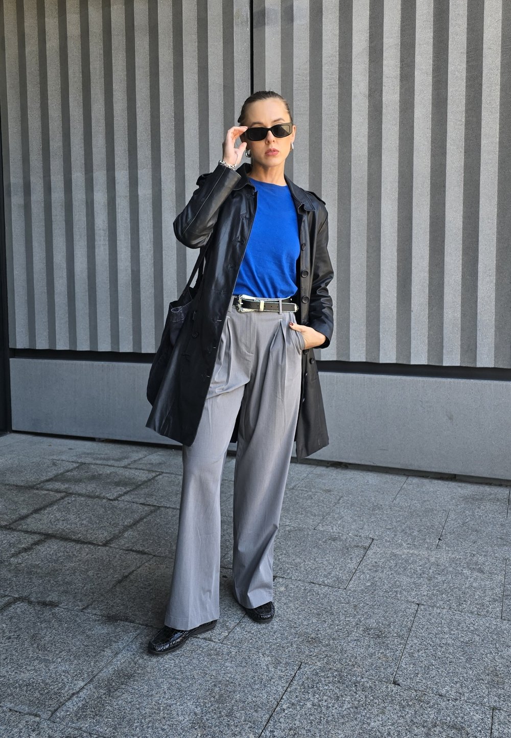 Μαύρο δερμάτινο παλτό πάνω από μπλε πουκάμισο, γκρι πλισέ παντελόνι και γυαλιστερά μαύρα παπούτσια. Τα αξεσουάρ περιλαμβάνουν γυαλιά ηλίου και μαύρη ζώνη.