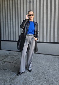 Μαύρο δερμάτινο παλτό πάνω από μπλε πουκάμισο, γκρι πλισέ παντελόνι και γυαλιστερά μαύρα παπούτσια. Τα αξεσουάρ περιλαμβάνουν γυαλιά ηλίου και μαύρη ζώνη.