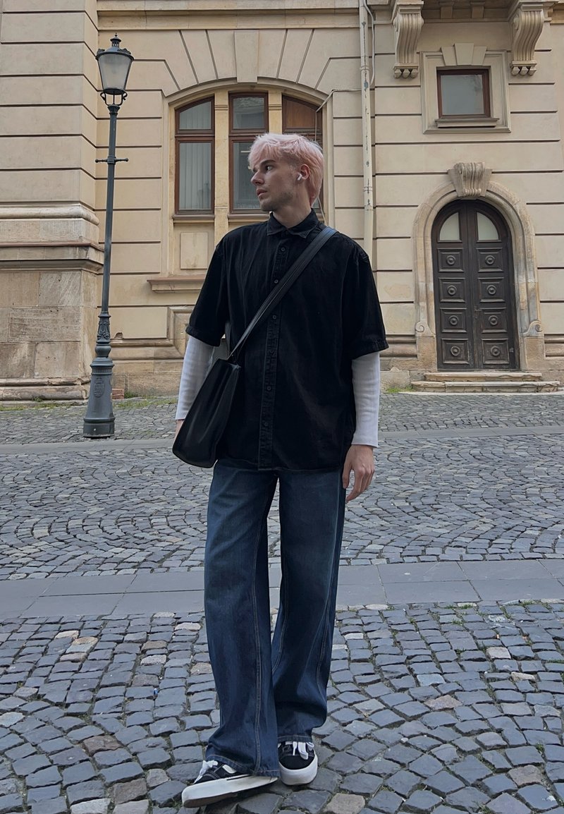 Jeune personne aux cheveux roses portant un chemisier noir, un sac et un jean large se tenant sur une rue pavée près d’un bâtiment historique et d’un réverbère.