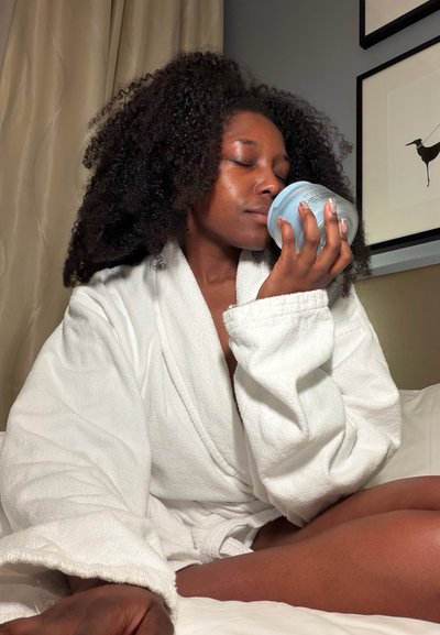 Mujer con bata blanca sentada en la cama, ojos cerrados, oliendo un tarro azul de crema o producto para el cuidado de la piel.