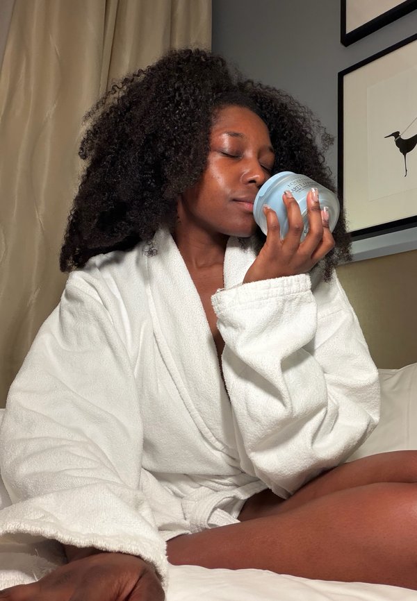 Donna in accappatoio bianco seduta sul letto, occhi chiusi, che annusa un barattolo blu di crema o prodotto per la cura della pelle.
