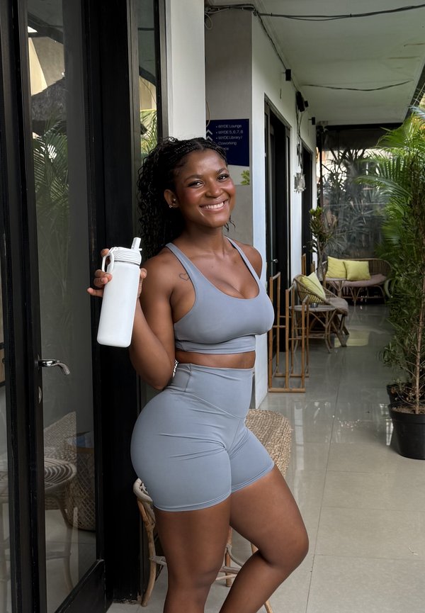 Lachende vrouw in grijze sportkleding houdt een witte waterfles vast, staand in een lichte hal met planten en rotanmeubilair.