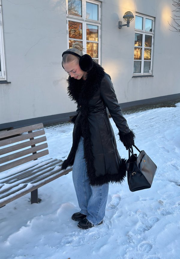 Vrouw in een zwarte jas met bontkraag, oorwarmers, handschoenen en spijkerbroek staat op sneeuw bij een houten bankje buiten een gebouw met ramen.