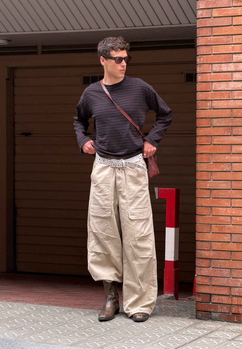 Homme portant des lunettes de soleil, une chemise noire à rayures à manches longues, un pantalon cargo beige large, une ceinture blanche, des bottes marron et un sac bandoulière, debout près d'un mur en briques.