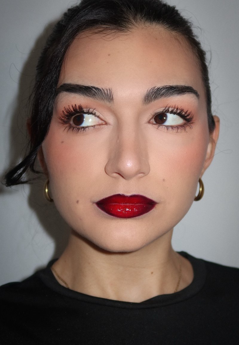 Femme aux cheveux foncés, portant un rouge à lèvres rouge brillant audacieux, de longs cils, des boucles d'oreilles en or en forme d'anneau, regardant vers sa gauche sur un fond uni.