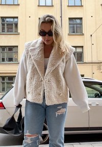 Creme faux fur vest med krave, dobbeltknaplukning; parret med en hvid langærmet top og slidte blå jeans. Sort håndtaske.