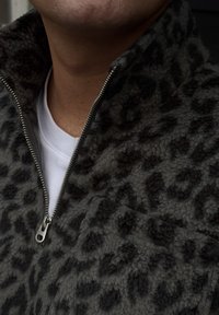 Flisasta jakna s kapuco v temno sivem odtenku z vzorcem leopardjega tiska. Ima visok ovratnik in srebrno zadrgo na sprednji strani.