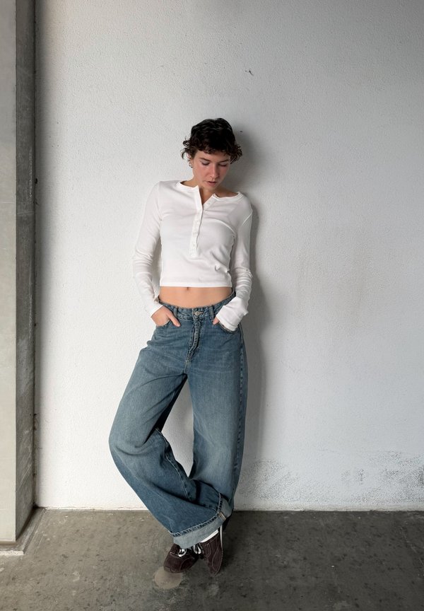 Person with short curly hair wearing a white cropped long-sleeve top and loose blue jeans, standing against a plain white wall.