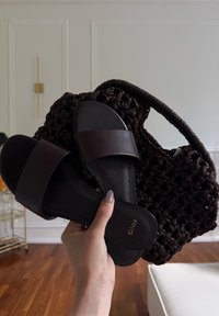 Mano sosteniendo sandalias planas negras tipo slide y un bolso tejido marrón oscuro en una habitación con suelo de madera y paredes blancas.