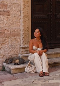 Beige trägerloses Oberteil, weite cremefarbene Hose, braune Sandalen und eine gehäkelte Tasche. Eine graue getigerte Katze ruht in der Nähe auf Steinstufen.