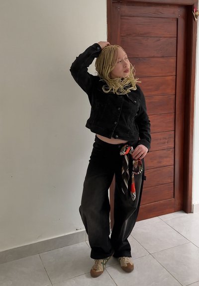 Chaqueta de mezclilla negra, diseño corto; pantalones negros de corte holgado con una bufanda colorida atada a la cintura; deportivas beige y verdes.