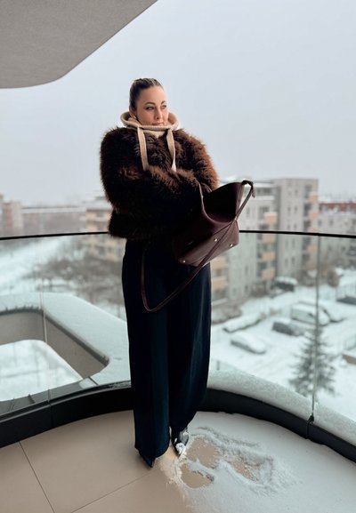 Chaqueta de piel sintética marrón, capucha beige, pantalones anchos negros, sosteniendo un bolso marrón oscuro con detalles metálicos, de pie en un balcón nevado.