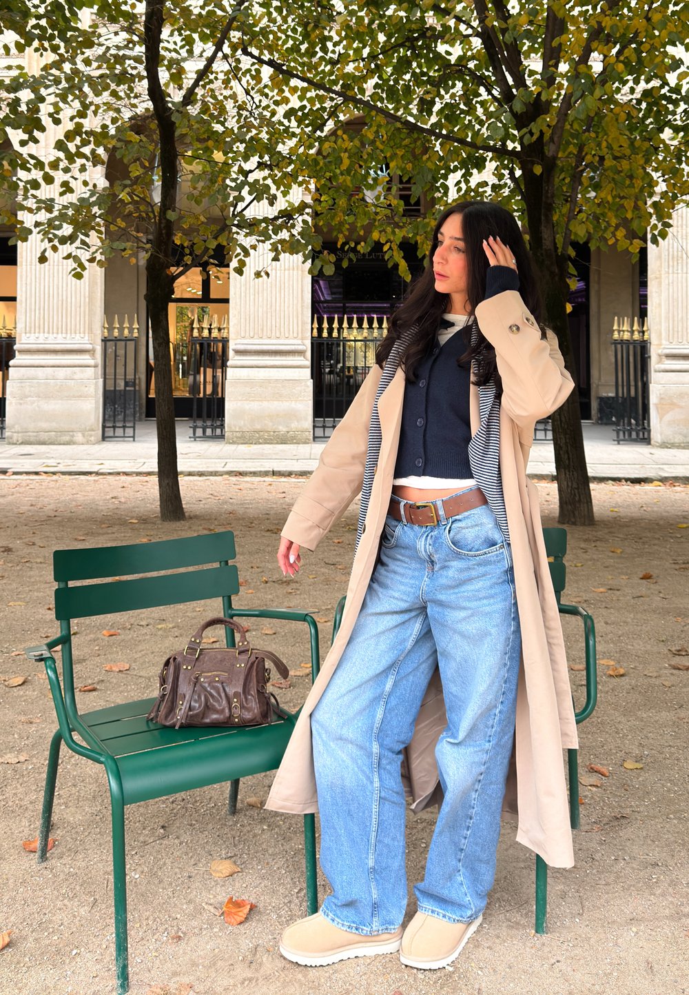 Beige trench coat, tengerészen pulóver, széles lábú kék farmer, barna slip-on cipő. Barna táska zöld széken, háttérben oszlopok és fák láthatók.