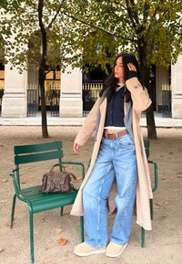 Beige trench coat, tengerészen pulóver, széles lábú kék farmer, barna slip-on cipő. Barna táska zöld széken, háttérben oszlopok és fák láthatók.