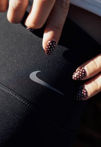 Leggings esportivos pretos apresentam um destacado logótipo Nike Swoosh cinzento. Uma mão com unhas castanhas com padrão de bolinhas aponta para a superfície das leggings.