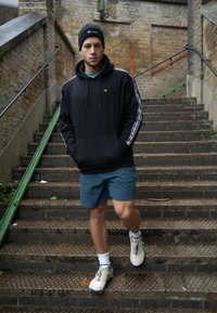 Sudadera con capucha negra y rayas blancas en los laterales, pantalones cortos turquesa y zapatillas ligeras. Modelo bajando escaleras de piedra con barandilla verde.
