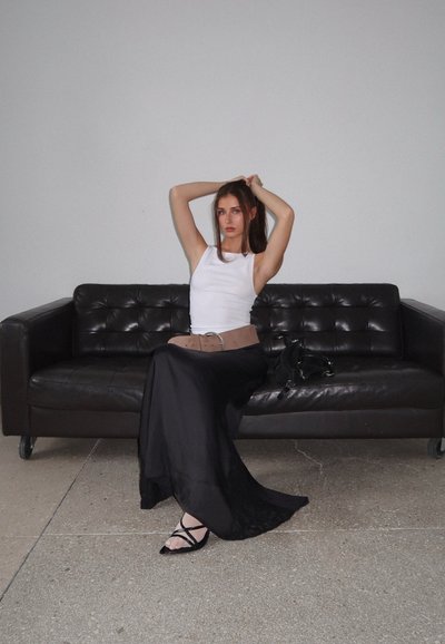 Falda maxi de satén negra con borde de encaje, camiseta blanca ajustada, cintura con cinturón marrón, sandalias negras de tiras, sentada en un sofá de cuero oscuro.