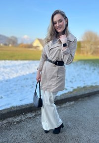 Bežinė tranšėja su juodu diržu, derinama su baltomis plačiomis kelnėmis. Juodas rankinės ir aukštakulnių batai. Fone matomas sniegas ir žolė.