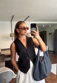 Γυναίκα με μαλλιά πιασμένα πίσω, φορώντας κόκκινες γυαλιά ηλίου, μαύρο γιλέκο, λευκή φούστα και κρατώντας μεγάλη μπλε τσάντα από σκοινί, που βγάζει selfie σε εσωτερικό χώρο.