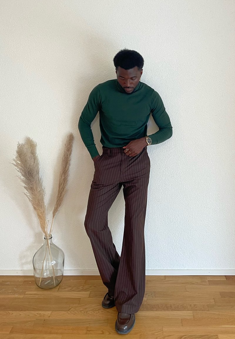 Chemise verte à manches longues, pantalon large à rayures brunes, et chaussures foncées. Vase en verre avec de l'herbe séchée en arrière-plan sur un sol en bois.