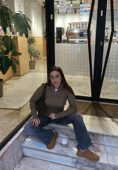 Suéter marrón de cuello con cremallera y logo "MNG", jeans azules y botas de slip-on beige. Sosteniendo una pequeña taza de café, sentado en escaleras de mármol. Entorno interior.