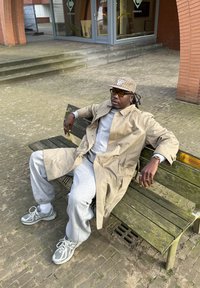 Beige trenchcoat över grå sweatshirt och sweatpants, sittande på en träbänk. Grå atletiska skor och en mönstrad keps med gula tonade glasögon.